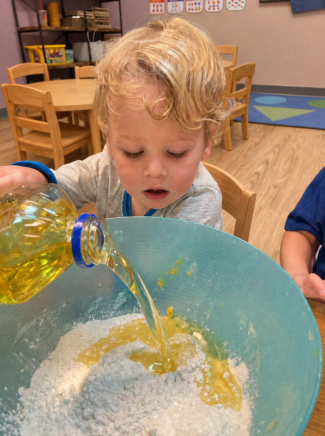 A child pouring liquid into a large blue mixing bowl during a classroom activity at L&L Lakeview.